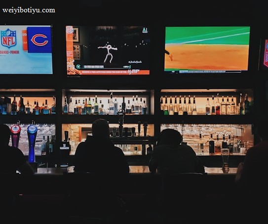 A bar counter with several people sitting in front of it is shown. Above the counter, there are multiple television screens displaying sports broadcasts, including football and baseball. Behind the counter, various bottles of alcohol are neatly arranged.