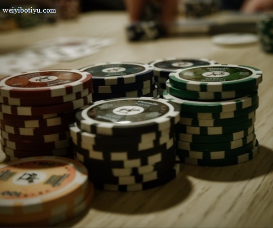 Stacks of poker chips are arranged on a wooden table, with various denominations and colors such as red, green, and black. Some playing cards are blurred in the background, indicating a game setting.