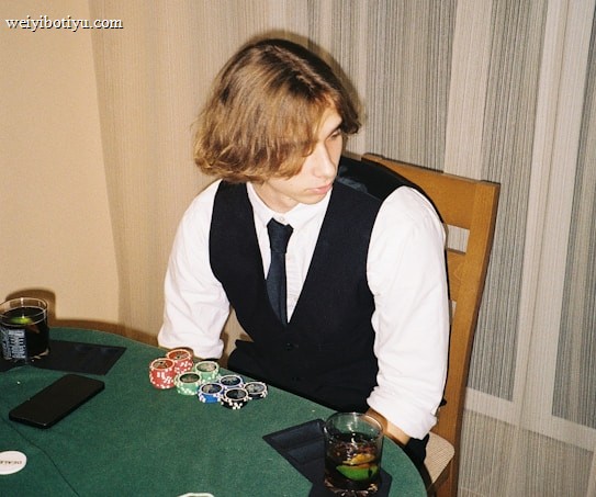 A person with shoulder-length hair is sitting at a poker table. They are wearing a white shirt, black vest, and tie, with an array of poker chips in front of them. There are two glasses with drinks on the table, and a smartphone is placed nearby. The background consists of vertical striped curtains.